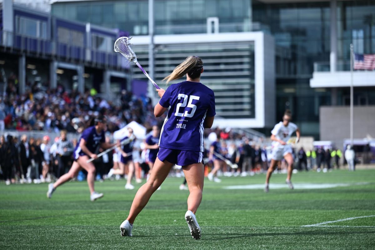 A purple-clad lacrosse player faces a full crowd in the stands.