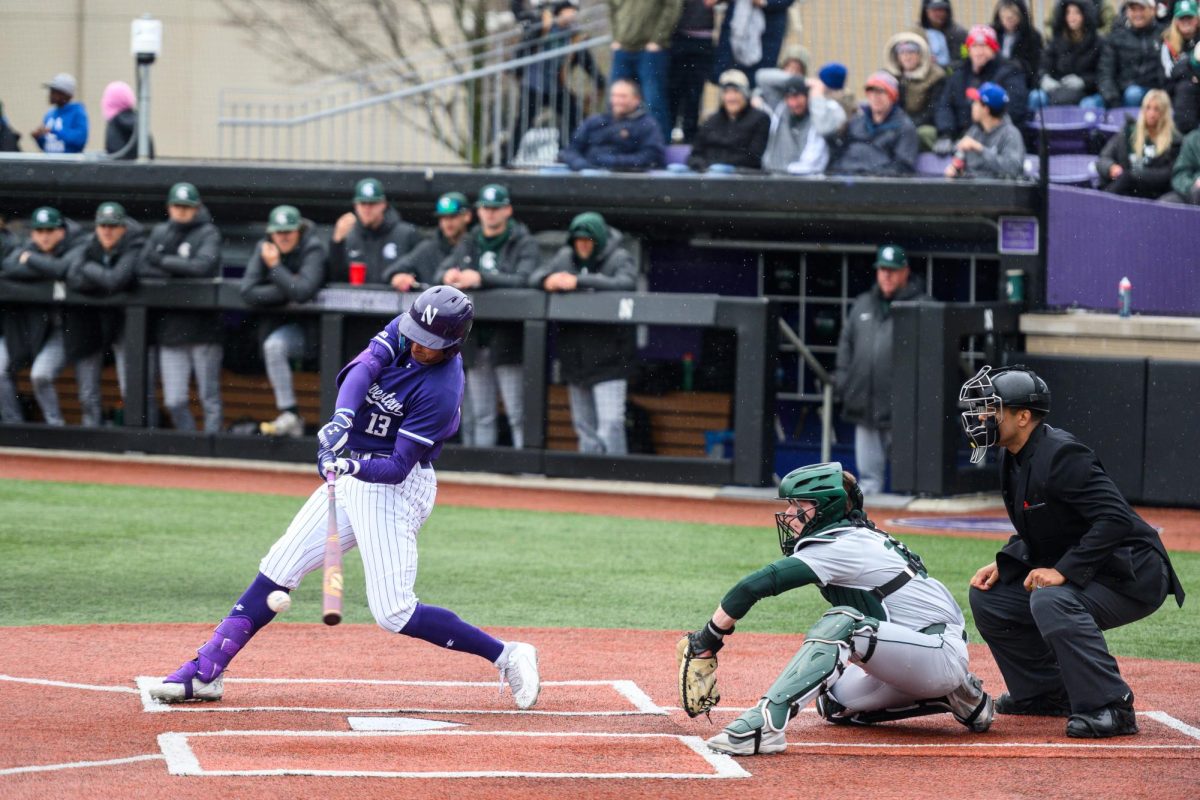 A man in purple swings a bat at a baseball as a man in green and gray sits behind.
