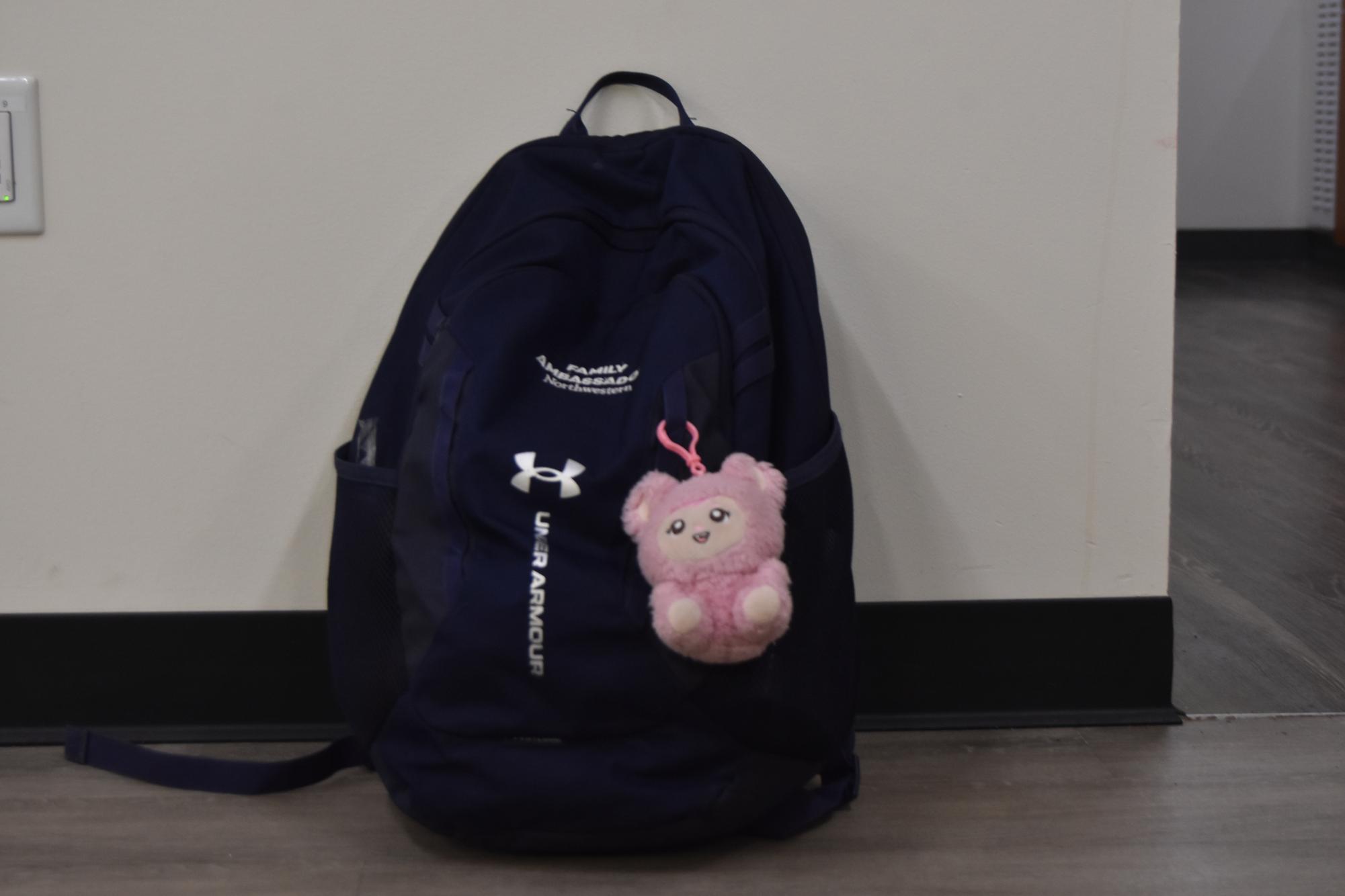 A blue bag with a pink keychain and silver lettering lays against a white wall.