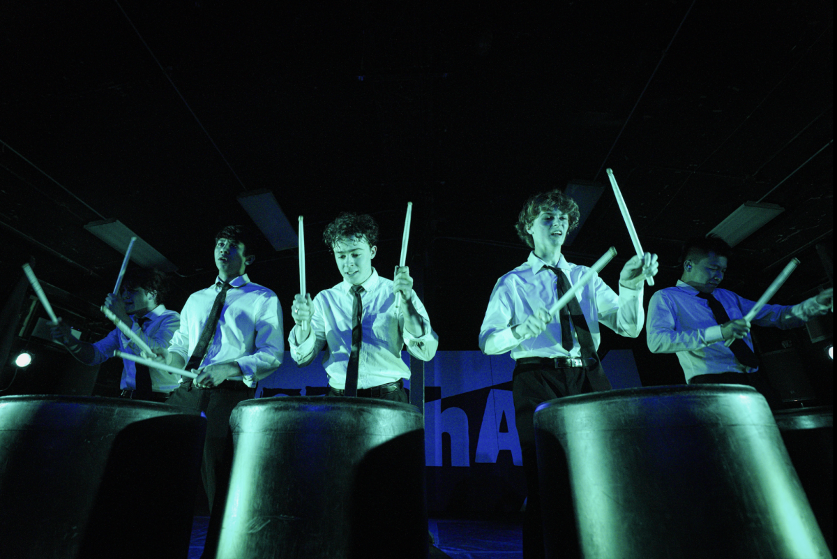 Drummers drum on black bins under green lighting.