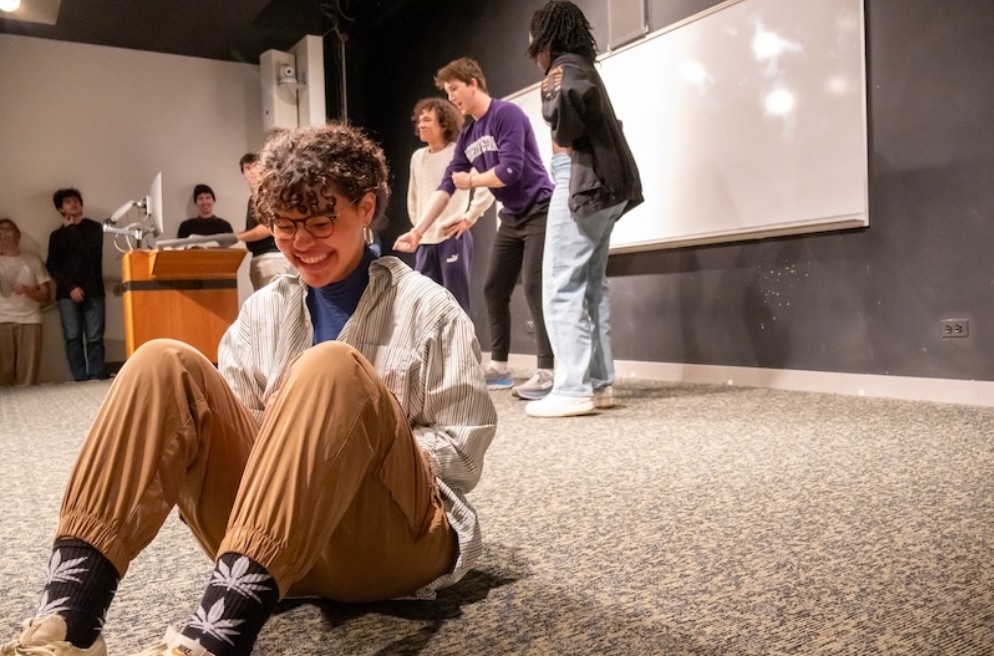 A performer sits on the front of the stage, reacting to the movements of the three actors behind her.