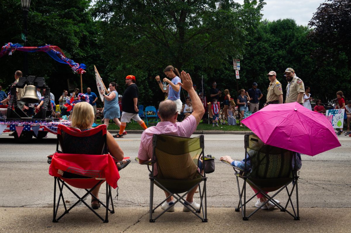Three people sit in chairs and watch a parade.