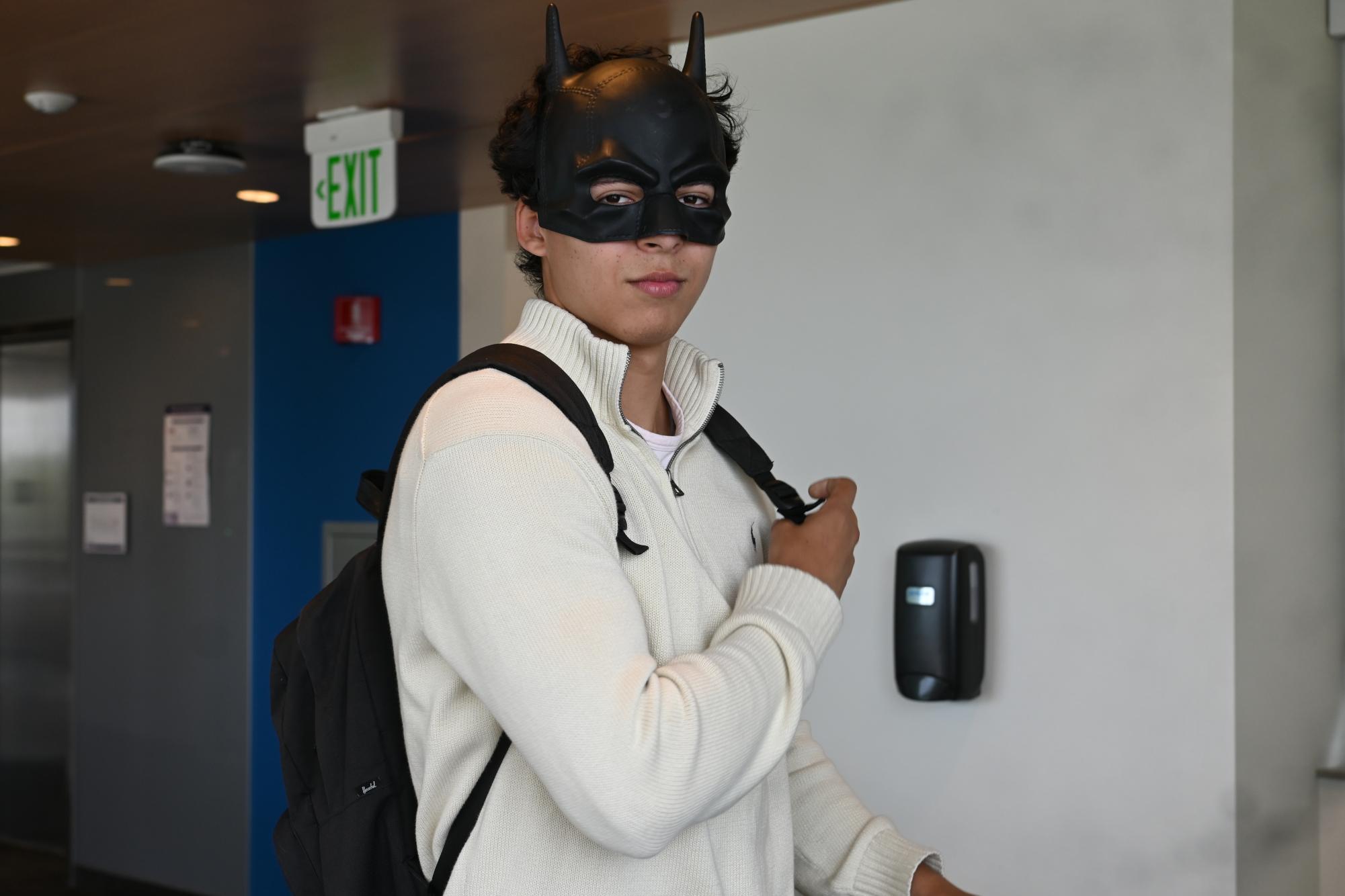 A young man poses wearing a black Batman mask holding the strap of a black backpack.