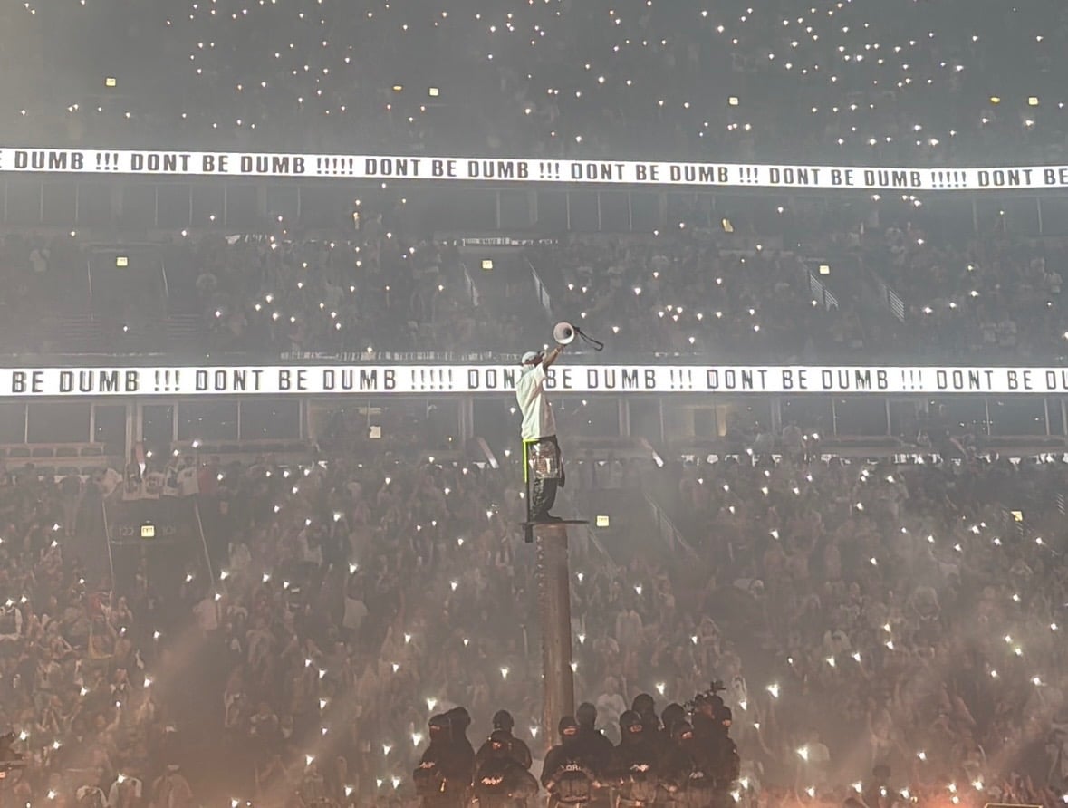 A person stands on an elevated platform with their hands up lifting a megaphone in front of a huge crowd.
