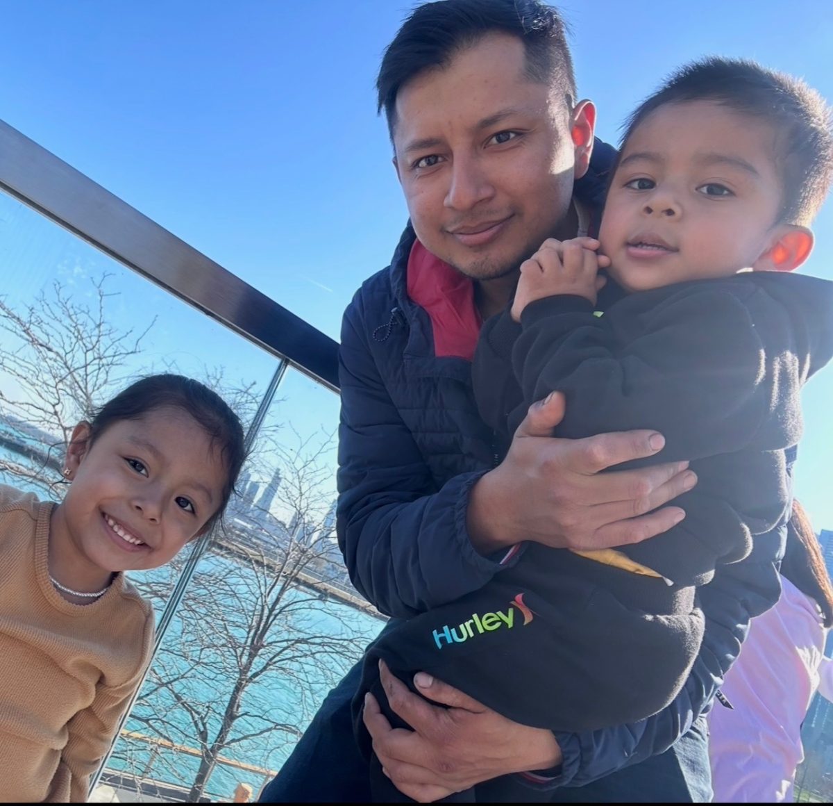 An adult holds a child in his arms, smiling, as another child smiles at the camera.