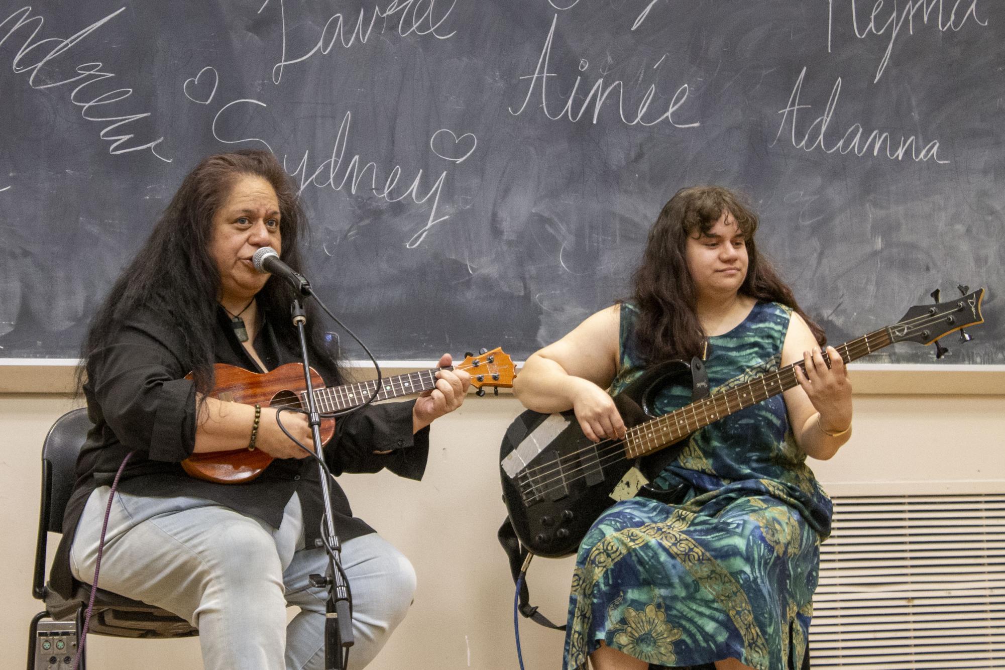 One performer sings into a microphone and plays ukulele as another performer plays guitar.