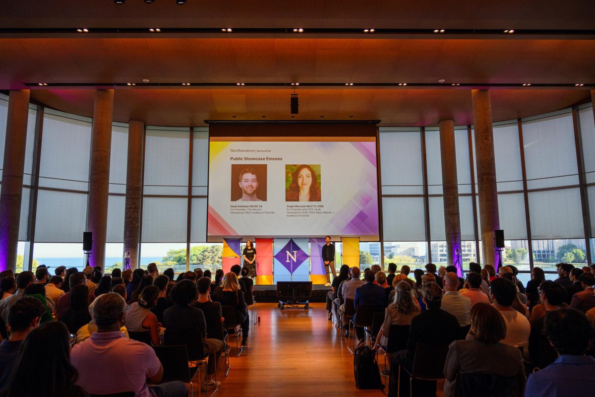 An audience watches a presentation on stage with large windows behind them.