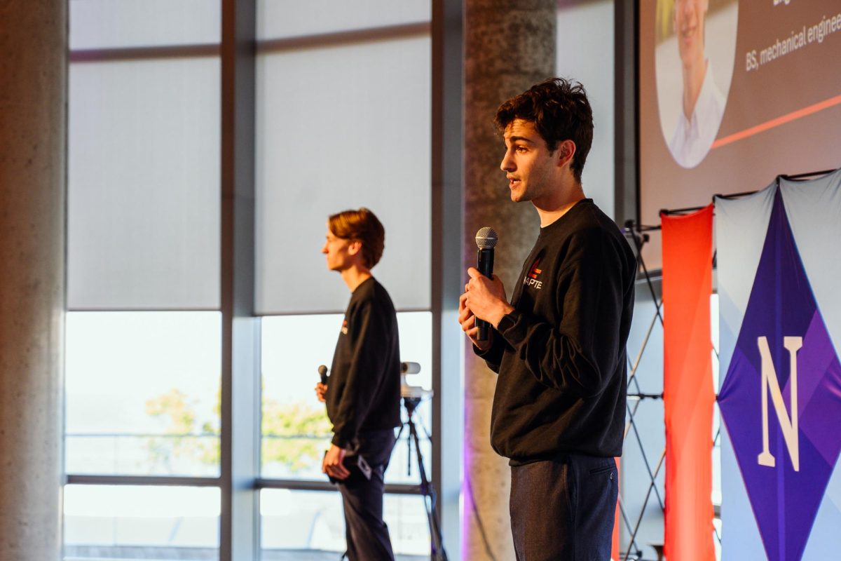 A person wearing black speaks into a microphone as another person in black stands next to them.