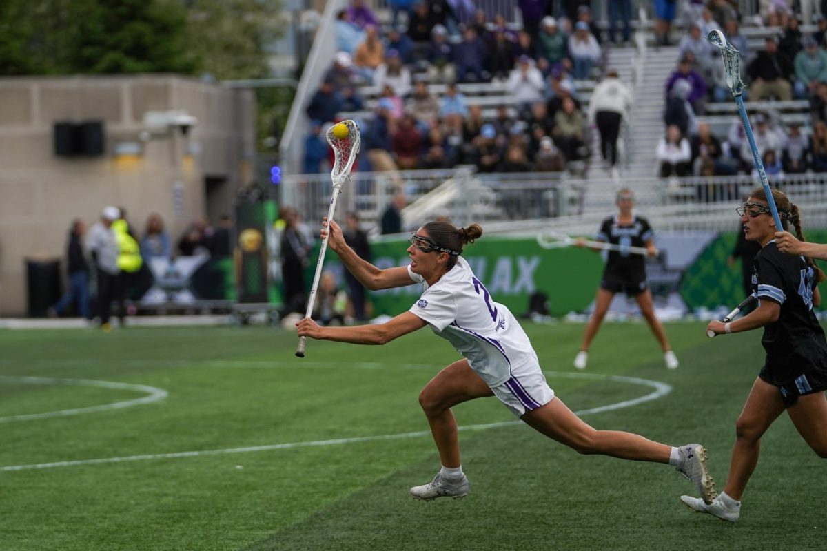 A player in white holds a ball in her stick while running down a field.