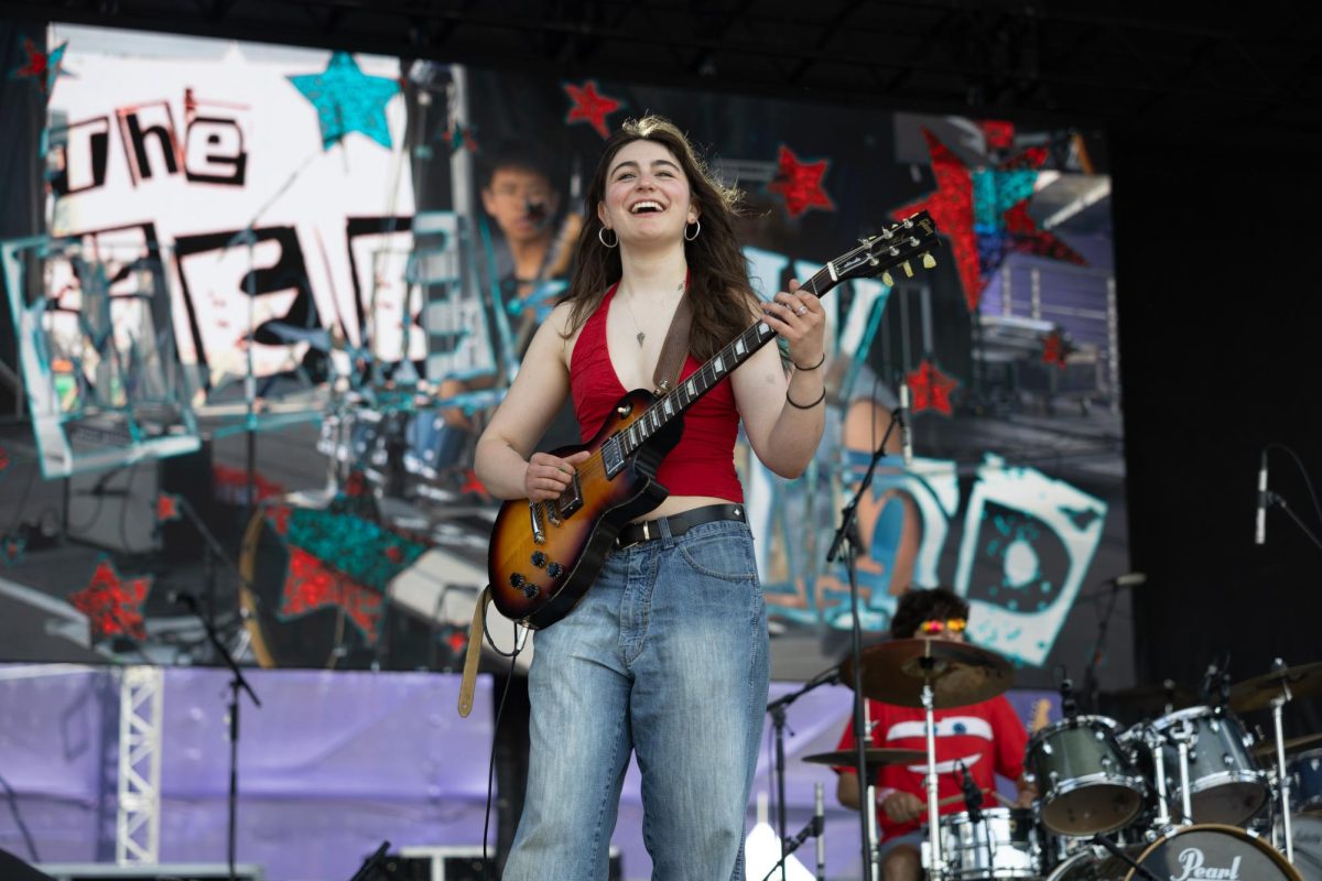 A performer wearing red plays a guitar and smiles broadly.