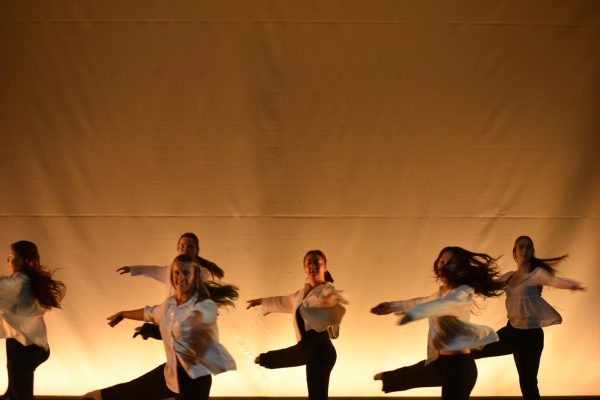 Six dancers face the camera while turning. Their arms are parallel and one leg is outstretched.