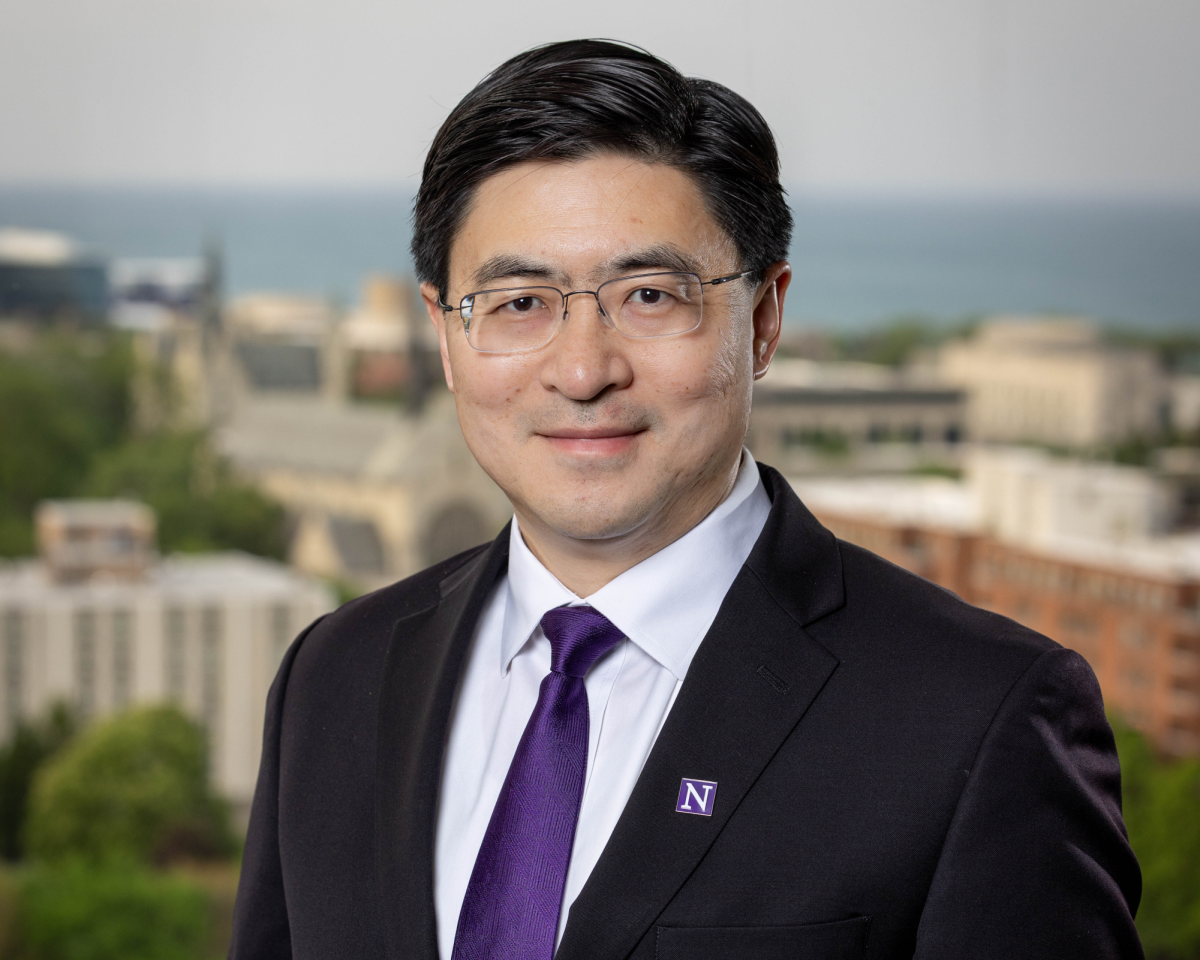 Una persona con un traje puesto con una corbata morada y una calcomanía del logotipo de Northwestern posa para una foto en frente de un fondo borroso del campus de Evanston de Northwestern.