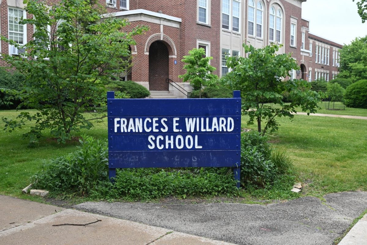 A blue sign with white lettering that reads “FRANCES E. WILLARD SCHOOL” in front of a brick school building with greenery.