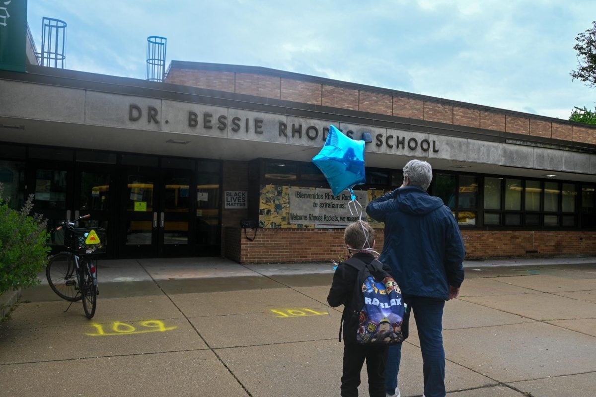 A child and an adult stand in front of a building with the text “DR. BESSIE RHODES SCHOOL” on it.
