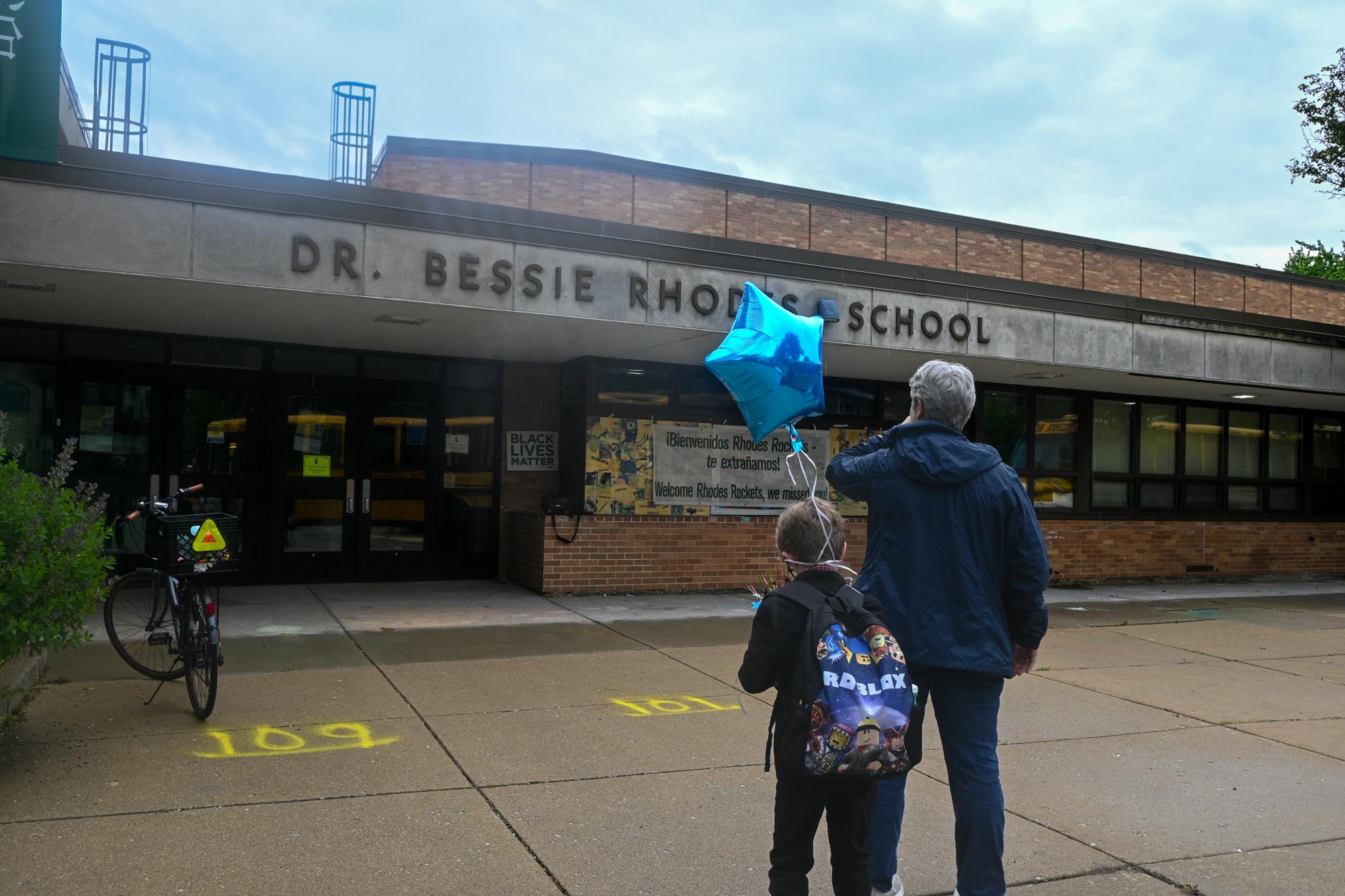 A child and an adult stand in front of a building with the text “DR. BESSIE RHODES SCHOOL” on it.