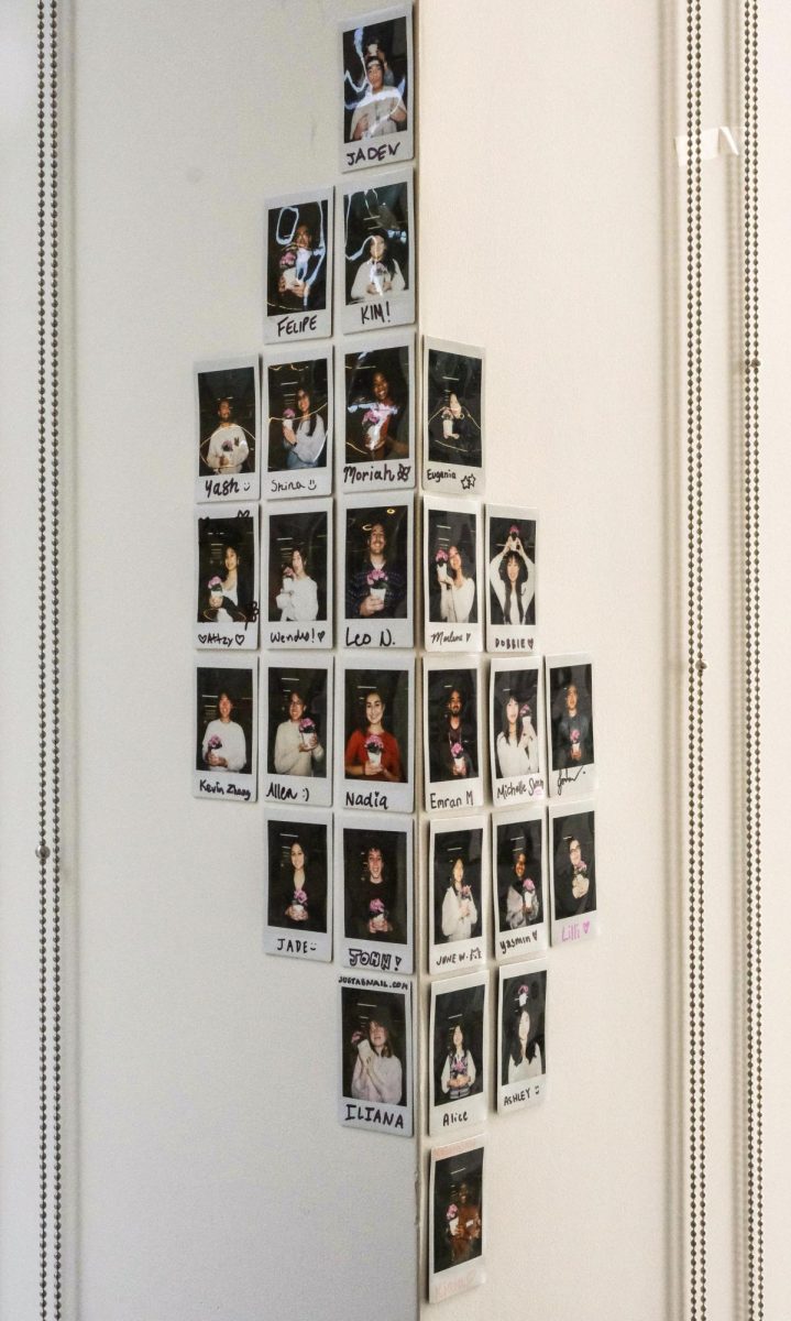 27 individual Polaroid photos with hand-written names underneath are arranged in a diamond pattern across the corner of two white walls.