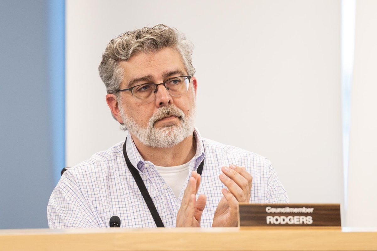 A person claps with a neutral facial expression. A placard on the desk in front of them says “Councilmember Rodgers.”