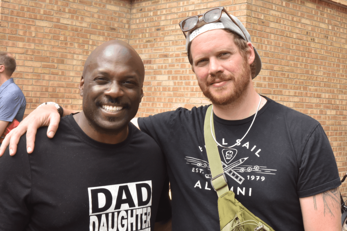 Two men in black shirts stand in front of a brick building.