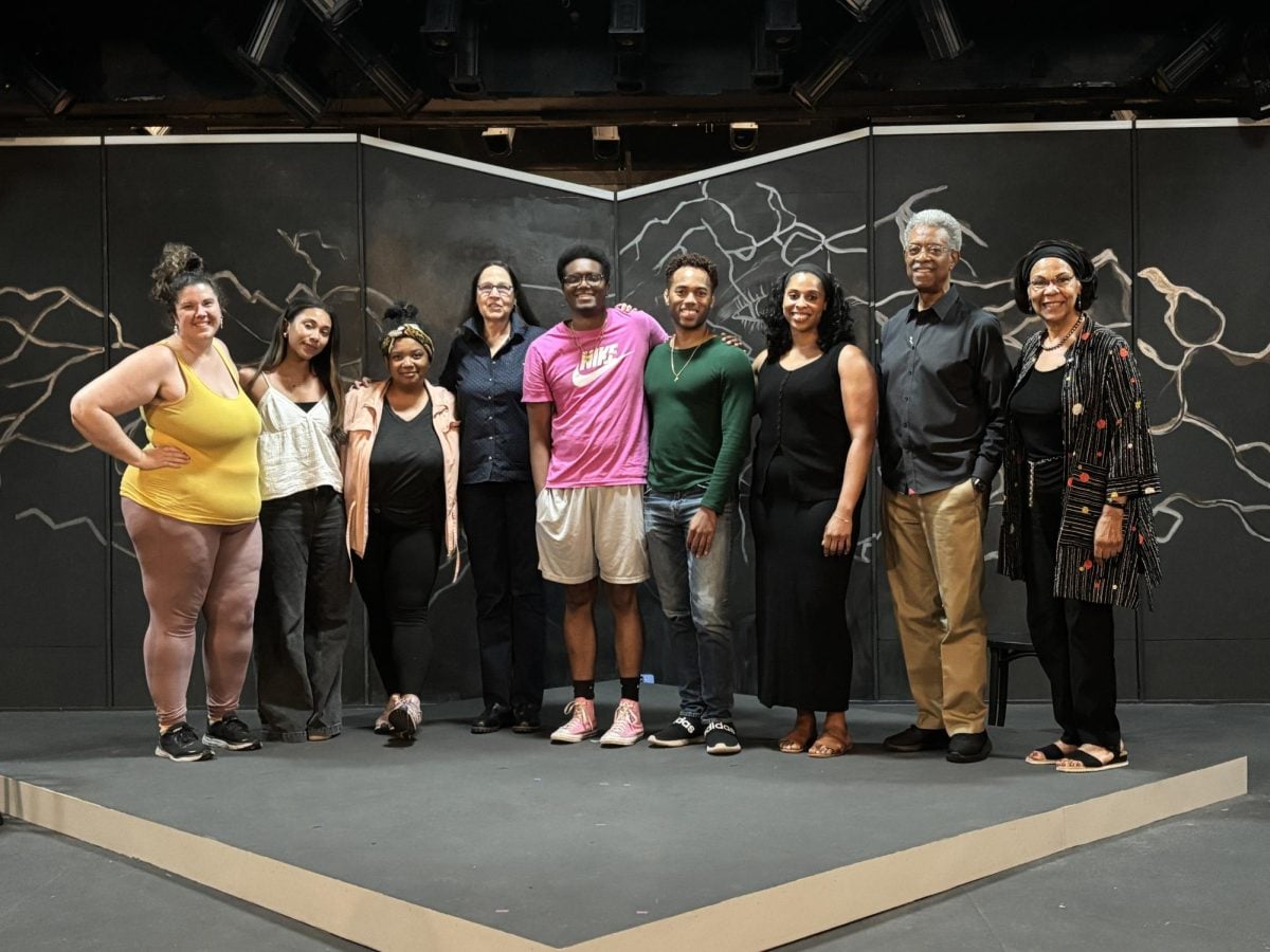 The cast and crew for a play reading poses for a group photo on a stage.
