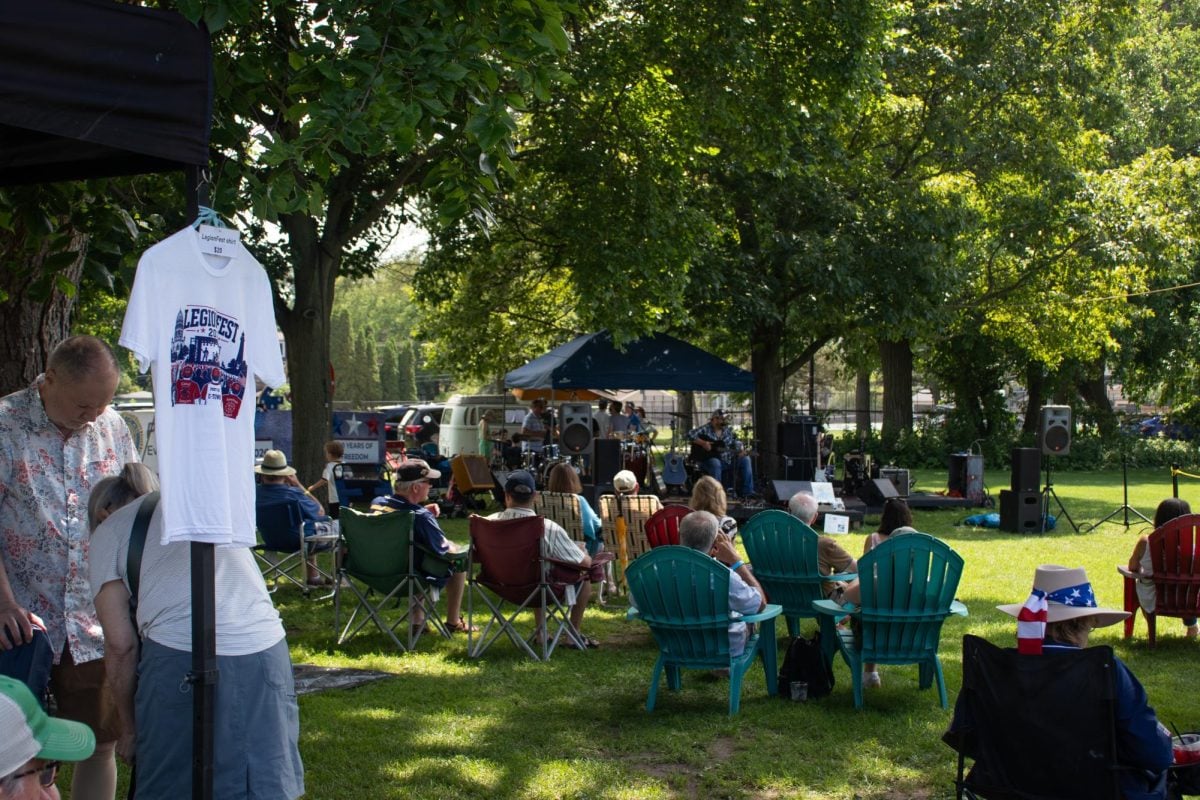 People sit on lawn chairs and blankets in the grass watching an outdoor concert.