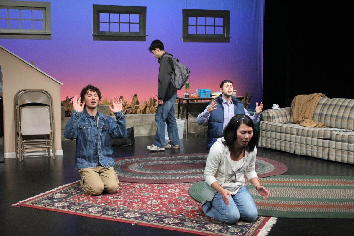 Three people kneel on the ground praying, with a fourth person standing behind them.