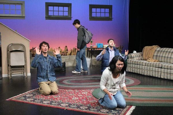 Three people kneel on the ground praying, with a fourth person standing behind them.