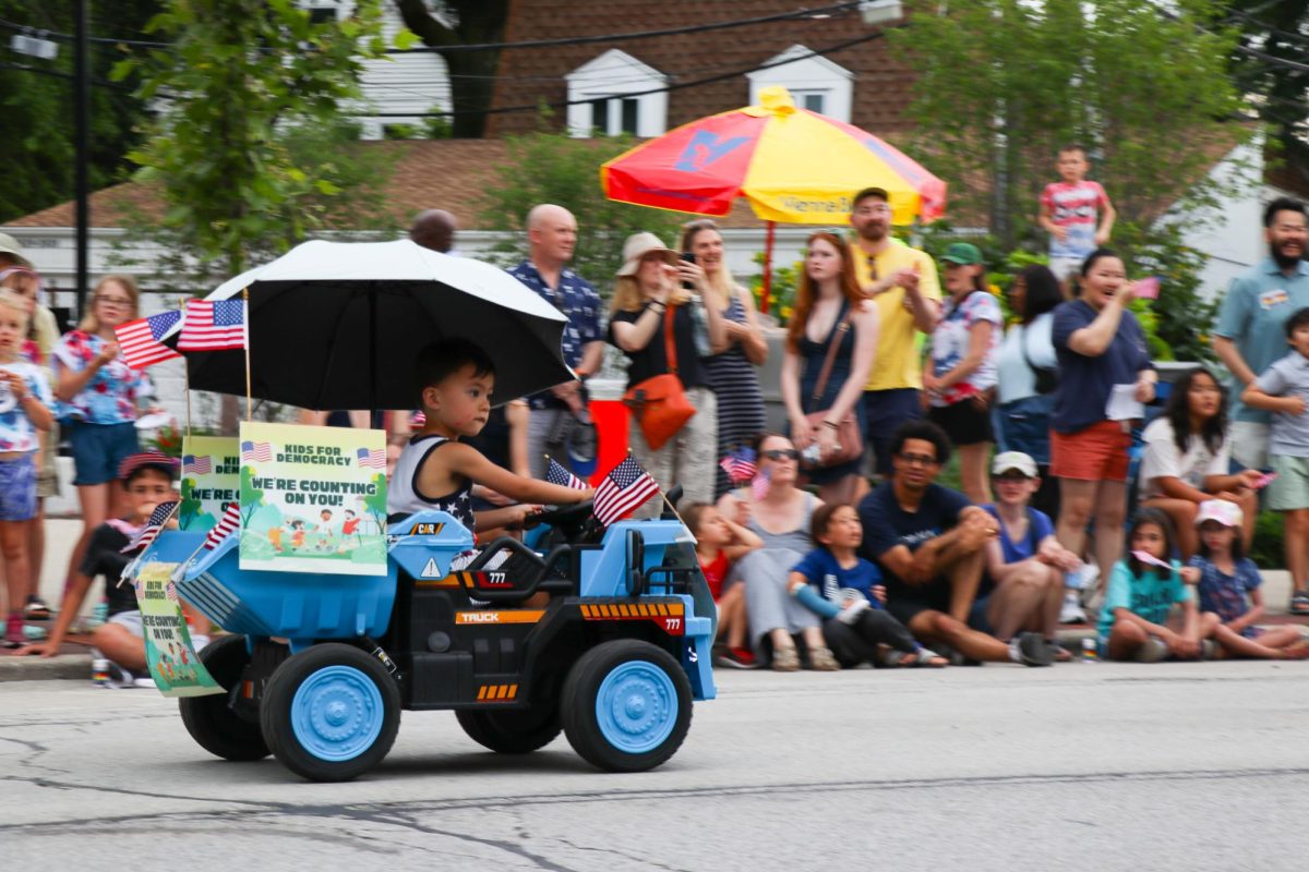 A child in a blue truck flying American flags drives to the right.