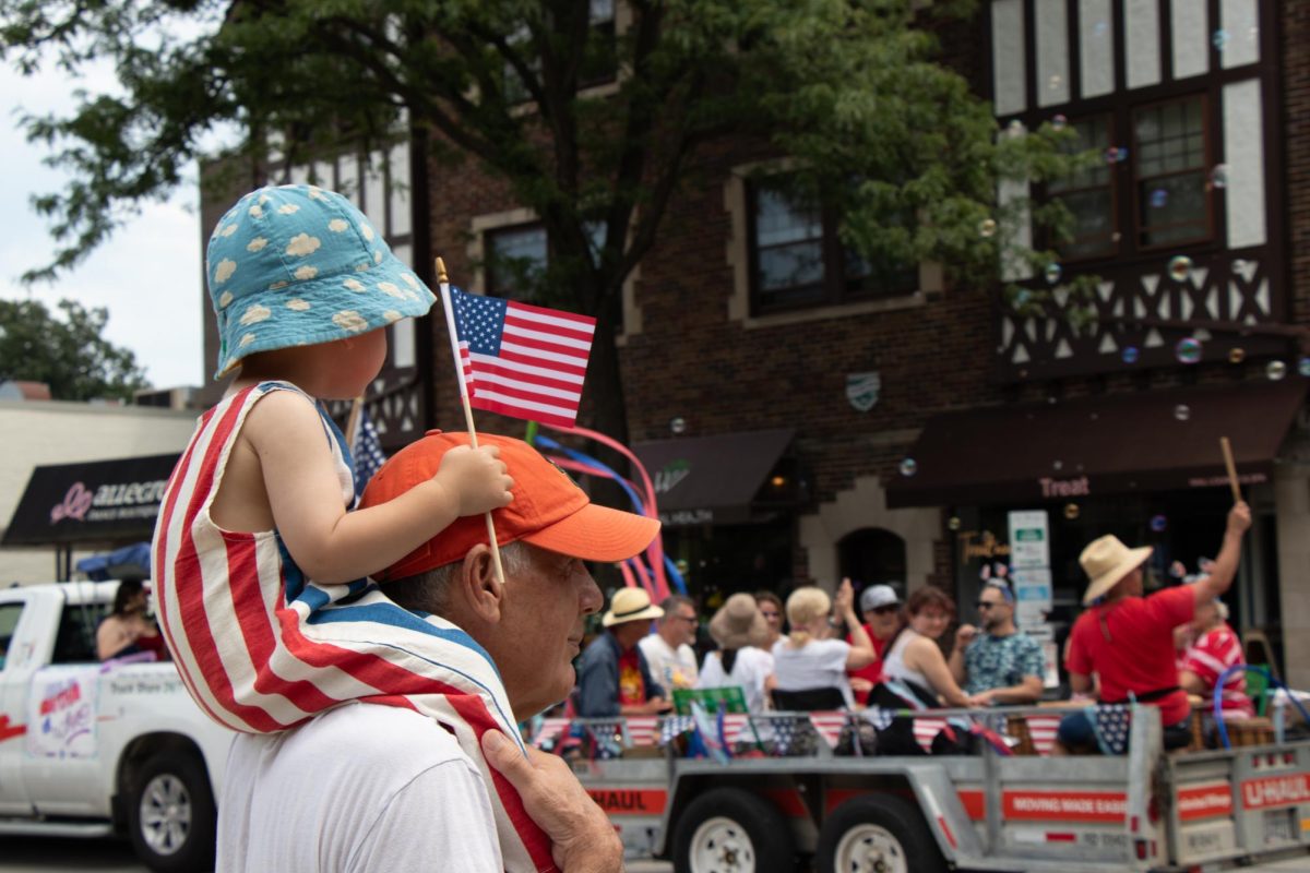 A child holds a mini American flag, sitting on a person’s shoulders