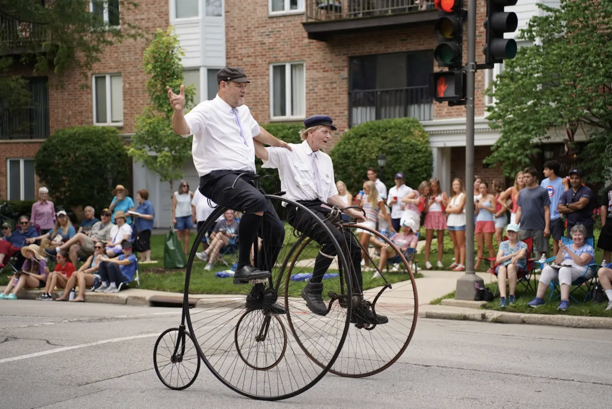 Two bikers ride penny-farthings, holding onto each other’s shoulders