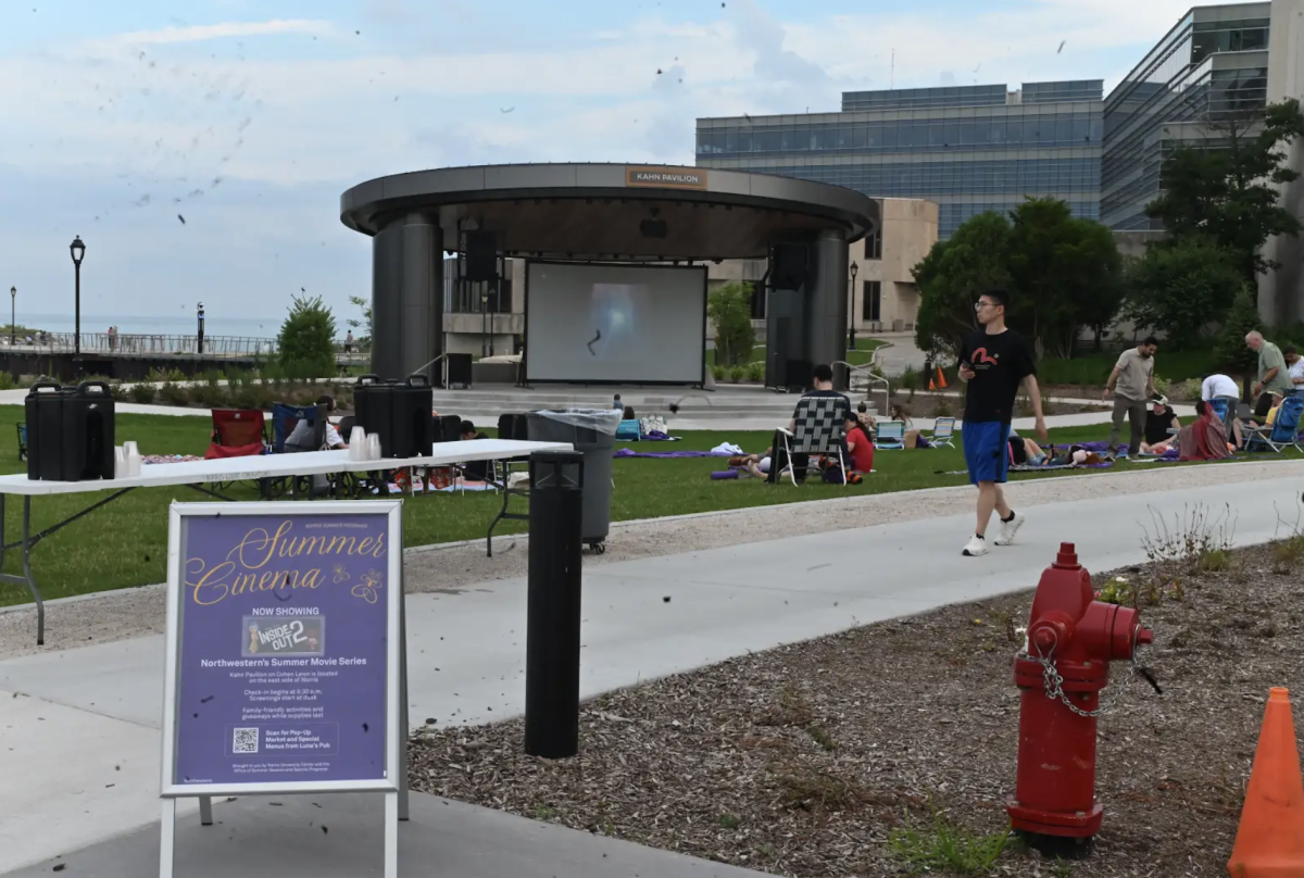 A sign that reads “Summer Cinema” is posted on a lawn where people sit on blankets and chairs.